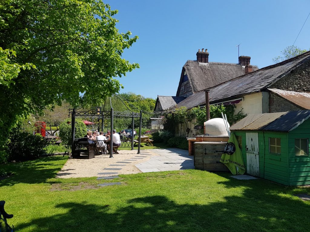 A peaceful outdoor dining area at The Golden Swan, Wilcot, featuring a shaded seating space under a metal pergola. The surrounding garden is lush with greenery, and a traditional thatched roof building sits in the background. The scene captures a sunny day with guests enjoying their time outside amidst natural beauty. A small green shed and outdoor wood-fired oven add to the rustic charm of the setting.