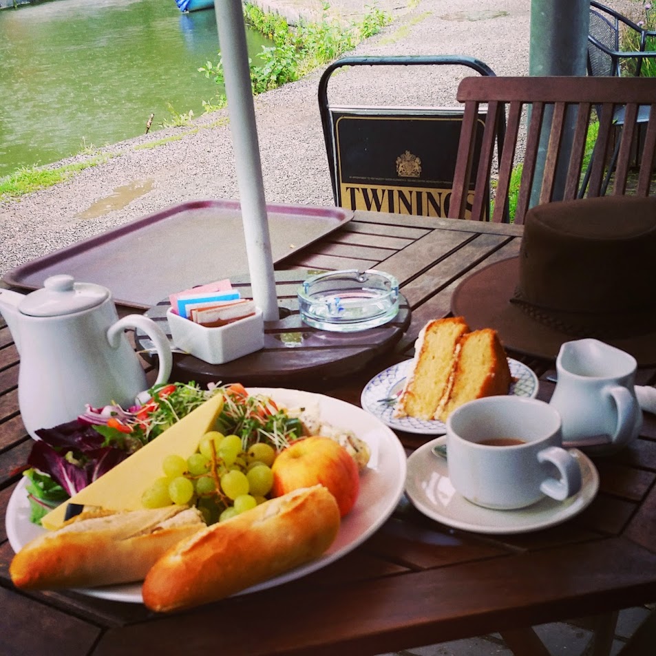 The image shows a delightful Ploughman’s lunch served at an outdoor café table by the water. The meal includes: A wedge of cheddar cheese. A small baguette, halved. Fresh green grapes and an apple. A mixed salad with greens and shredded vegetables. A small serving of coleslaw. Beside the plate are a pot of tea, a teacup with milk jug, a slice of sponge cake on a separate plate, and a hat resting on the table. The peaceful riverside setting creates a serene and cozy atmosphere, perfect for enjoying a relaxed lunch.