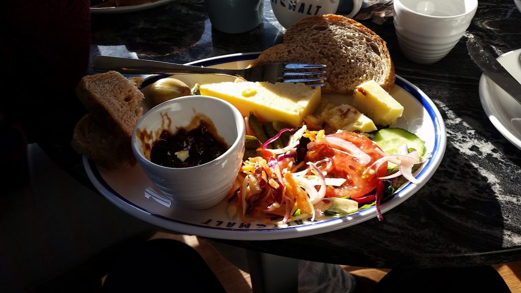 The image shows a Ploughman’s lunch served on a plate from "Salt and Malt." The meal features several components: Thick slices of cheddar cheese. A small dish of chutney or relish. A mixed salad with tomato, cucumber, red onion, and shredded vegetables. A few slices of hearty wholegrain bread, stacked beside pickles and a pickled onion. Some gherkins are also visible alongside the salad. This meal is a classic Ploughman’s style lunch, with a balance of cheese, bread, and fresh salad, complemented by the sweet and tangy chutney. Sunlight highlights the plate, creating a warm and inviting atmosphere.