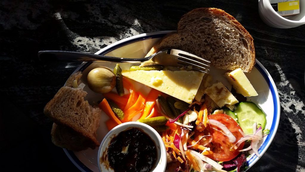 
The image shows a flat view of a Ploughman’s lunch, served on a plate from "Salt and Malt." The meal includes:

A wedge of cheddar cheese.
Slices of hearty wholegrain bread.
Pickled vegetables, including a whole pickled onion and gherkins.
Carrot sticks alongside the pickles.
A fresh salad with sliced cucumber, tomato, red onion, and shredded vegetables.
A small dish of dark chutney or relish.
The plate is bathed in sunlight, casting warm highlights on the fresh, colourful ingredients, creating an inviting, rustic feel.