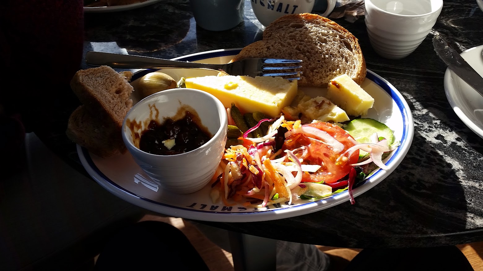 The image shows a Ploughman’s lunch served on a plate from "Salt and Malt." The meal features several components: Thick slices of cheddar cheese. A small dish of chutney or relish. A mixed salad with tomato, cucumber, red onion, and shredded vegetables. A few slices of hearty wholegrain bread, stacked beside pickles and a pickled onion. Some gherkins are also visible alongside the salad. This meal is a classic Ploughman’s style lunch, with a balance of cheese, bread, and fresh salad, complemented by the sweet and tangy chutney. Sunlight highlights the plate, creating a warm and inviting atmosphere.