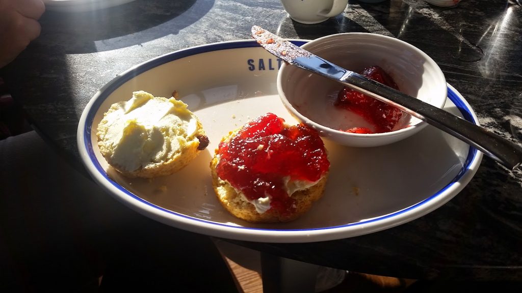 
The image shows a classic cream tea setup, likely from a café. On a plate labeled "Salt and Malt," there are two halves of a scone. One half is topped with a generous amount of clotted cream, and the other with both clotted cream and a thick layer of bright red strawberry jam. Next to the scone is a small bowl containing more jam, with a knife resting across it. Sunlight is streaming across the table, highlighting the rich textures of the scone and toppings, creating a warm and inviting scene for an afternoon tea.