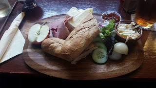 A wooden serving board holds a traditional Ploughman's lunch featuring a whole wheat baguette, slices of ham, cheddar cheese, a half apple, pickled onions, cucumber slices, mixed salad greens, and a small dish of coleslaw. A portion of chutney is placed alongside, completing the hearty meal. A pint of beer is visible in the background, set on a wooden table at The Lamb Inn, Axbridge.
