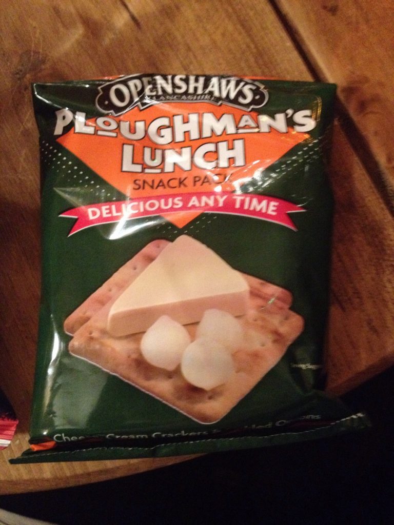 A close-up image of a snack pack labeled "Openshaws Ploughman's Lunch," featuring a green, orange, and black design. The slogan "Delicious Any Time" is prominently displayed on a red banner below the product name. The packaging suggests a convenient, ready-to-eat version of the traditional Ploughman's lunch, offering an easy snack option.