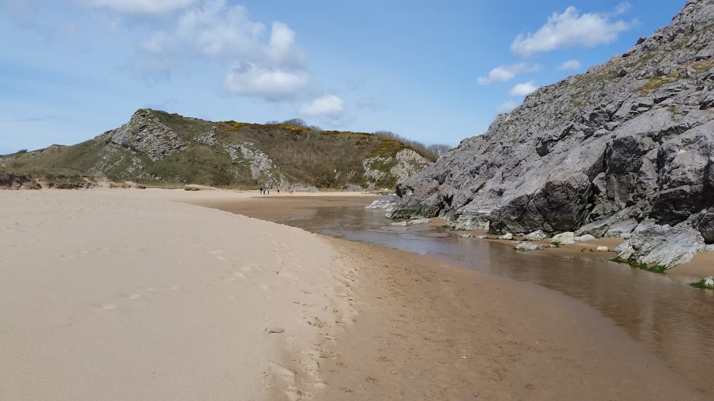 The image shows a scenic beach scene, likely taken at Bosherton Broad Haven Beach. The foreground is a wide expanse of sandy shore with gentle footprints leading toward the distance. A small stream or water channel runs alongside the sand, bordered by rugged rock formations on the right side. In the background, there are rolling grassy hills with patches of rocks and shrubs. A few people can be seen walking in the distance, adding a sense of scale to the vastness of the beach. The sky is blue with scattered white clouds, suggesting a clear, pleasant day at the beach.