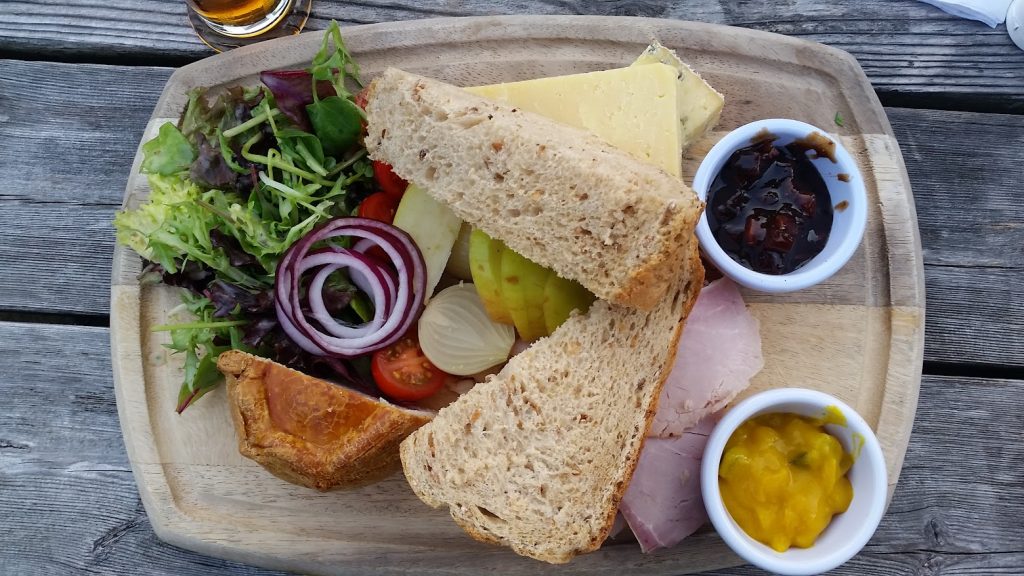 A flat-lay of a Ploughman's lunch at the Mayfly in Fullerton. The meal is served on a wooden board, featuring thick slices of multigrain bread, cheddar cheese, and generous portions of ham. A mini pork pie adds to the hearty presentation, with accompaniments of Branston pickle and bright yellow mustard piccalilli in separate ramekins. A fresh salad of mixed greens, sliced red onions, cherry tomatoes, and pickled onions completes the dish. The rustic wooden table and natural light enhance the inviting, countryside feel of the meal.