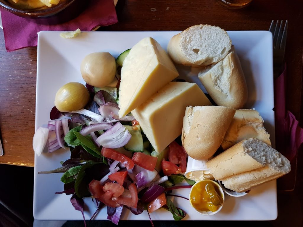 A flat-lay view of a Ploughman’s lunch served at the Boat Inn in Cromford. The meal is arranged on a large white square plate, featuring generous wedges of cheddar cheese, two pickled onions, fresh baguette slices, and a side salad with leafy greens, diced tomatoes, cucumbers, and red onion. Small pots of chutney and mustard add to the meal, with the bread and cheese providing the main focus. The plate is vibrant with colorful vegetables and a well-balanced presentation of classic Ploughman's elements. The rustic table and cozy pub atmosphere add to the charm.