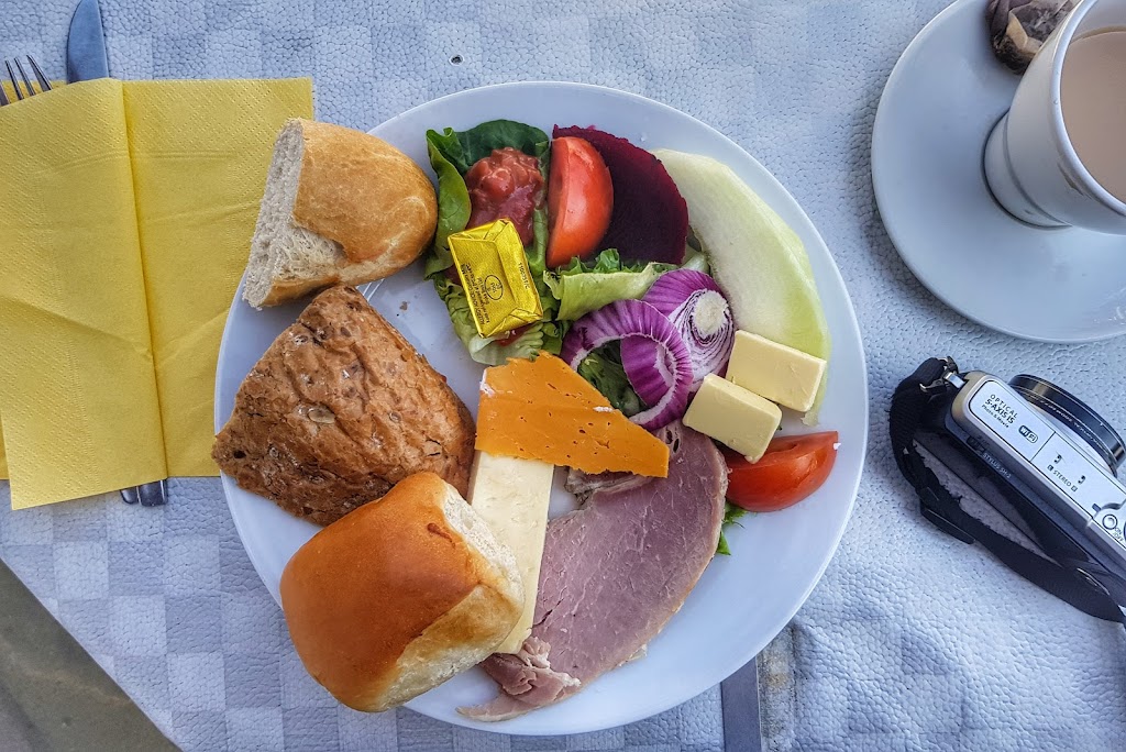A vibrant Ploughman’s lunch served on a white plate, featuring a variety of fresh ingredients. The plate includes a selection of cheeses like cheddar and soft cheese, slices of ham, a mix of salad greens with tomatoes, red onion, and beets, and a couple of different bread rolls. A small pat of butter is placed alongside the salad. A cup of tea and a camera are placed nearby, indicating an outdoor dining experience. The napkins are yellow, adding a bright contrast to the scene.