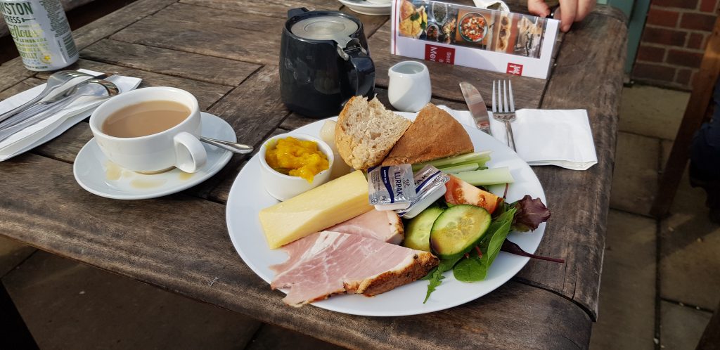 The image shows a classic Ploughman’s lunch served on a white plate. The meal includes thick slices of ham and cheddar cheese, a rustic bread roll, celery sticks, sliced cucumber, and a mixed salad with lettuce and tomato. There's a small dish of bright yellow piccalilli and packets of Lurpak butter resting on the cheese. To the side, there's a cup of tea, partly filled, on a white saucer, along with a teapot nearby. The table is wooden, and utensils like a knife, fork, and spoon are arranged neatly, with a menu booklet partially visible in the background. The setting suggests an outdoor, relaxed dining environment.