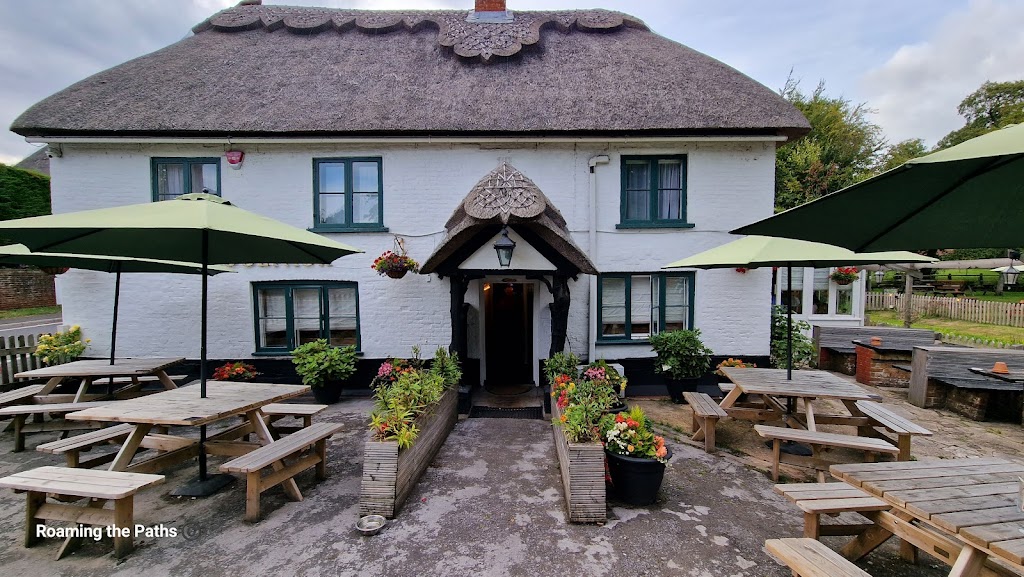 The image shows the front exterior of The Woolpack in Sopley, a charming traditional English pub with a white-painted facade and a distinctive thatched roof. The pub features a quaint, rustic entrance, framed by a decorative wooden arch. Bright flower arrangements in pots and planters line the walkway, adding vibrant color to the scene.

Two large green umbrellas cover wooden picnic benches set outside the pub, creating a welcoming outdoor seating area for patrons. The building's simple design, small windows with dark green frames, and wooden beams give it a cozy, countryside atmosphere. The patio area appears inviting and well-maintained, perfect for enjoying a casual meal or drink.