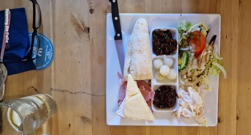
A flatlay view of a Ploughman’s lunch served on a square white plate. The meal includes a wedge of cheddar cheese and a baguette positioned diagonally, along with sliced ham. The center of the plate features a divided dish containing pickled onions, dark chutney, and cucumber slices. On the right side, there is a fresh salad of lettuce, tomato, cucumber, and onion drizzled with balsamic glaze, next to a creamy coleslaw. A knife rests alongside the baguette, and glasses and a pair of eyeglasses are visible in the background on a wooden table.