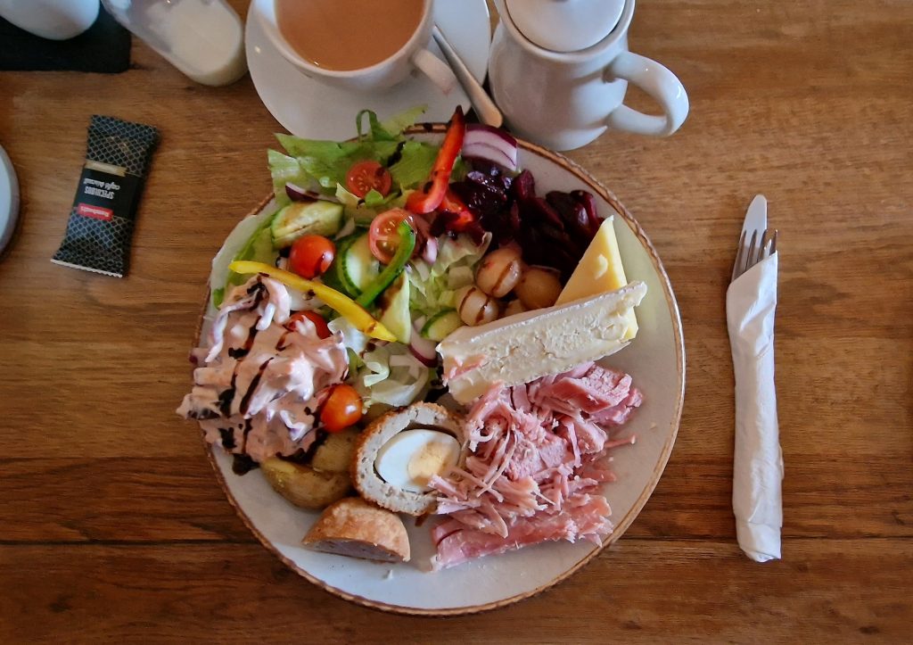 A flatlay of a generously portioned Ploughman's lunch served on a round plate. The meal includes a variety of classic components: thick slices of creamy Brie and mature Cheddar cheese, a pile of tender, shredded ham, and half a Scotch egg with its golden yolk exposed. Alongside the proteins, there are pickled onions, sliced beetroot, and a fresh salad with lettuce, cherry tomatoes, cucumber, red onion, and bell peppers, drizzled with a balsamic glaze. The salad also includes a creamy homemade coleslaw. In the foreground, a piece of pork pie is visible, adding a hearty, savory element. The plate sits on a wooden table, next to a neatly wrapped set of utensils and a cup of tea with milk. A teapot and milk jug are placed nearby, enhancing the traditional British meal setting.