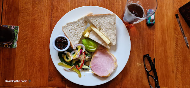 This image shows a flat-lay view of a classic Ploughman’s lunch served on a white plate, resting on a wooden table. The meal includes several traditional components arranged neatly across the plate. In the upper portion, two slices of plain white sandwich bread are stacked on top of each other. Tucked under the bread, there are two slices of cheddar cheese and a slice of brie cheese, creating a creamy contrast to the sandwich-style bread.
To the left, a small white ramekin holds a generous portion of dark chutney, which looks rich and tangy, ready to complement the cheese and bread. Beside the chutney is a fresh, colorful salad with mixed greens, sliced red onions, and strips of red and yellow bell peppers, lightly drizzled with a mustard dressing. The salad adds a vibrant, fresh element to the otherwise rich and hearty plate.
In the lower right corner, there is a thick slice of ham with a slightly pink center and well-defined edges, which appears to be home-cooked or freshly carved, contributing to the traditional feel of the meal. Next to the ham, there are slices of crisp green apple, adding a sweet and tangy balance to the dish.
In the background, you can see a half-filled pint glass of what appears to be ale, placed to the top right of the plate. A pair of black eyeglasses and a pen sit to the right of the plate, suggesting a relaxed pub setting. The rich brown, wooden texture of the table adds a rustic touch, enhancing the hearty, comforting nature of this traditional British pub meal. Additionally, a colorful drink coaster with a partial glass of a dark-colored beverage is visible to the left, further completing the atmosphere of a cozy dining experience.