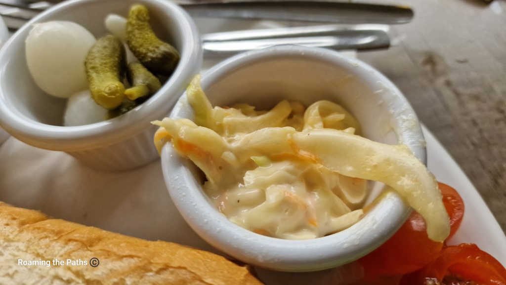The image shows a close-up view of two small ramekins on a plate, each containing classic ploughman's accompaniments. The ramekin on the left holds a few tiny pickled onions and small gherkins (cornichons), offering a crunchy and tangy bite. The ramekin on the right contains a creamy serving of freshly made coleslaw, with visible strands of cabbage and carrot coated in mayonnaise. In the background, part of a crusty baguette and halved cherry tomatoes are also visible, along with cutlery, suggesting a traditional British ploughman's lunch setting. The textures and colors emphasize the variety of flavors on the plate.