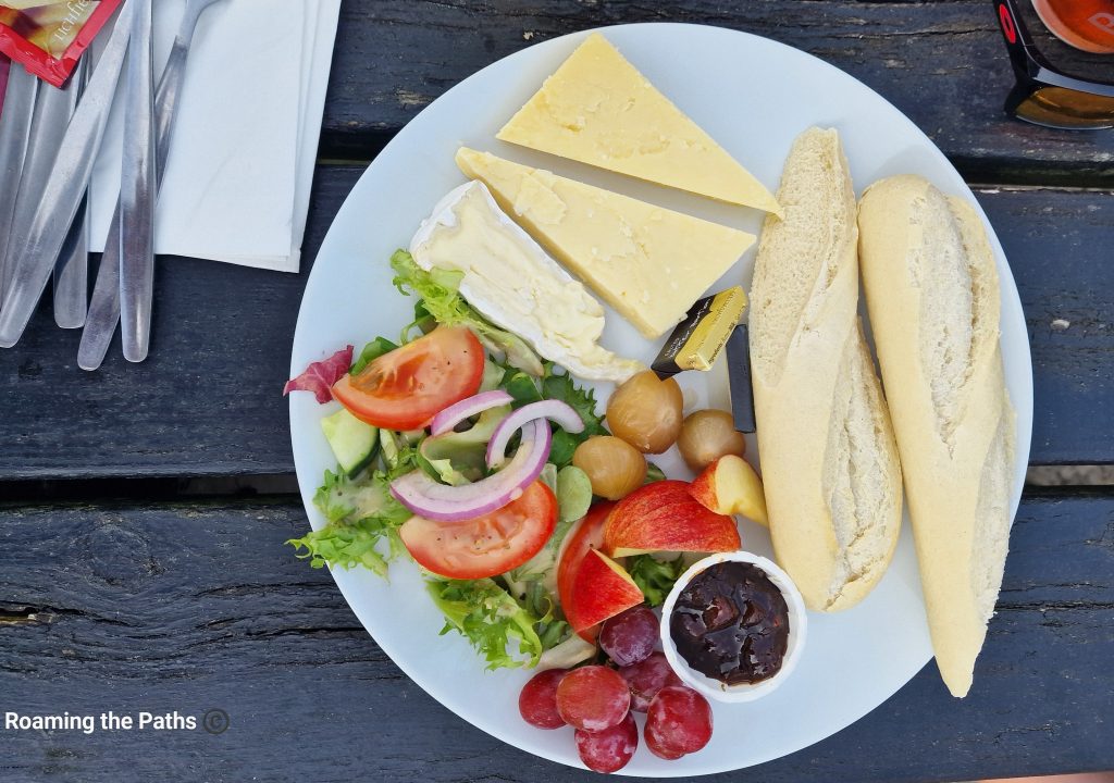 The image shows a traditional Ploughman’s lunch flatlay, elegantly arranged on a white plate set against a weathered wooden surface. The meal includes two thick slices of mature cheddar cheese and a wedge of soft, creamy Brie. A portion of crusty baguette is sliced lengthwise into two pieces, placed to the right of the cheeses.
On the left side of the plate is a fresh, vibrant salad consisting of crisp lettuce, juicy slices of tomato, thin rings of red onion, and cucumber, all lightly dressed. Nestled beside the salad are several small pickled onions, adding a tangy contrast to the dish.
In the lower part of the plate, fresh fruit provides a refreshing sweetness, with red apple slices and a handful of red grapes. A small ramekin of chutney accompanies the fruit, likely intended as a complement to the cheeses.
Also on the plate are two small packets of butter, suggesting the baguette can be enjoyed with a spread of creamy butter to enhance the overall flavor. The presentation is simple yet appetizing, evoking the rustic charm of an English countryside meal.
Off to the side, silver cutlery wrapped in napkins and a red-tinted object (possibly sunglasses) are visible, hinting at an outdoor dining experience. The entire composition conveys a hearty, fresh, and delicious Ploughman’s lunch perfect for a sunny day.