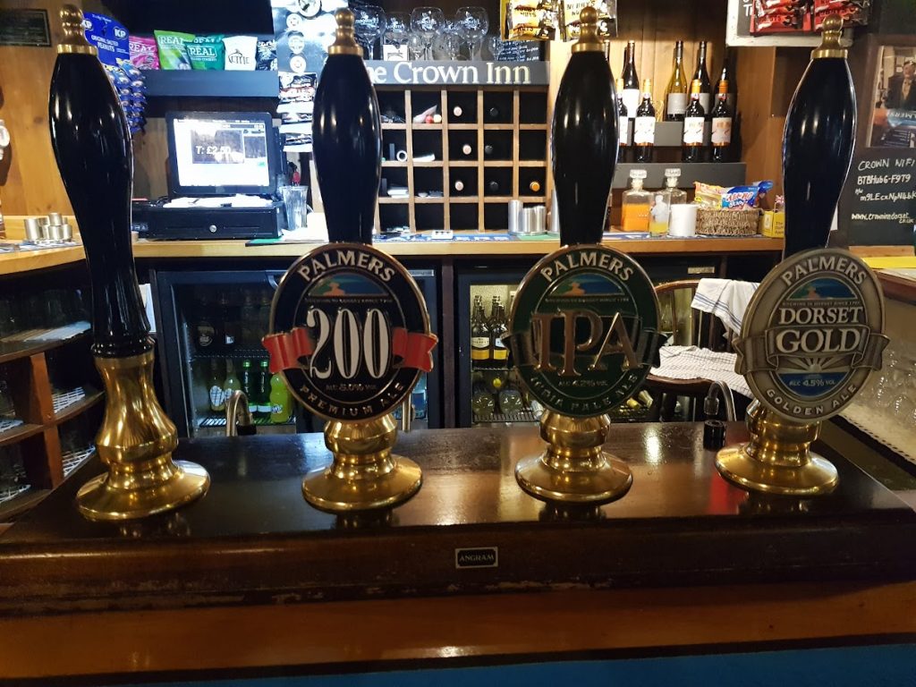 This image displays a close-up view of the bar at the Crown Inn, showcasing four traditional beer engine handles lined up neatly along the wooden countertop. The handles are used to serve local ales from Palmers Brewery, a well-known brewery in Bridport, Dorset. From left to right, the taps feature:
Palmers 200 Premium Ale: A dark label with "200" in bold red letters on a black background, indicating a premium ale.
Palmers IPA: The green label features the iconic IPA (India Pale Ale) designation.
Palmers Dorset Gold: A golden ale, highlighted with a light-colored label featuring "Dorset Gold."
The backdrop of the bar shows neatly arranged bottles of wine, various bar snacks, and a cash register. The overall atmosphere evokes a traditional, cozy pub ambiance with its wooden accents, making it an inviting spot for visitors to enjoy these local brews.
