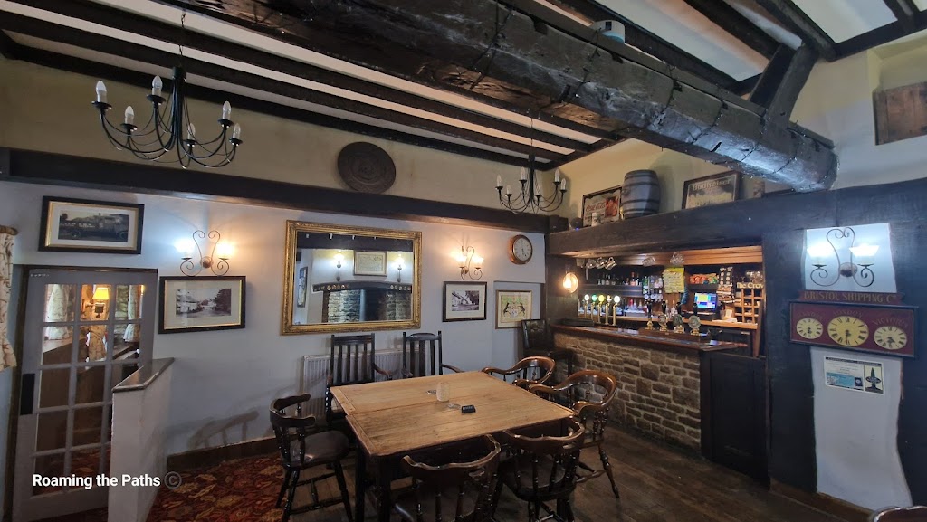 The image shows the cozy, rustic interior of the Crown Inn at Puncknowle, a traditional English country pub. The focal point is a large wooden table surrounded by dark wooden chairs with arms, providing an inviting spot for patrons to gather. Above, thick wooden ceiling beams span the room, adding a historic, rustic charm. Two black metal chandeliers with candle-style lights hang from the ceiling, further emphasizing the old-world ambiance.
In the background, the bar is neatly arranged with various beers on tap, highlighted by warm hanging lights. The bar front is made of stone, and a shelf behind it displays various bottles, glasses, and pub décor. On the wall to the left of the bar, vintage signs and framed paintings hang, while a large, golden-framed mirror reflects the room, adding depth. Above the bar, traditional pub signs and a decorative wooden barrel are displayed, giving the space a sense of heritage and authenticity.
A clock and framed artwork line the back walls, adding to the pub's homey feel. The carpet has a classic design, complementing the warm tones of the wood and stone. The overall atmosphere is inviting and reminiscent of a traditional English pub, with a sense of history and a welcoming vibe for visitors and locals alike.