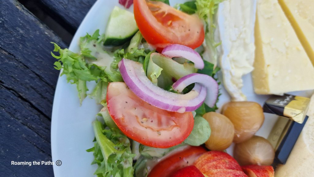 This image showcases a close-up of a fresh, vibrant salad from a Ploughman's lunch. The salad consists of crisp lettuce leaves, thick slices of juicy red tomatoes, and rings of sharp red onion. Accompanying these are chunks of cool cucumber, providing a refreshing crunch. The salad is lightly dressed with a vinaigrette, visible on the leaves and tomatoes, adding a slight sheen to the fresh produce.
To the right of the salad, there are small pickled onions, offering a tangy contrast to the fresh ingredients. The edge of the plate also features wedges of creamy Brie and mature cheddar, alongside wrapped butter pats. The colors of the salad — from the deep reds of the tomatoes and onions to the pale green of the lettuce — are set against the neutral tones of the cheese, creating a beautifully balanced, appetizing presentation. The salad appears fresh, crisp, and perfectly complements the rest of the meal. The rustic wooden table underneath adds a charming, countryside feel to the overall scene.