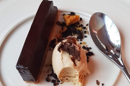 A gourmet dessert plated elegantly on a white dish. The dish features a sleek, rectangular slice of rich chocolate tart with a smooth, glossy finish. Next to it is a scoop of creamy vanilla or caramel ice cream, garnished with chocolate shavings. Scattered around the plate are small, textured crumbs, possibly cookie or brownie bits, along with a few pieces of honeycomb for added crunch. A reflective silver spoon is placed beside the dessert, hinting at the indulgent experience awaiting the diner. The composition of the plate highlights the contrast between the dark, decadent chocolate and the light, creamy ice cream, creating a visually appealing presentation.