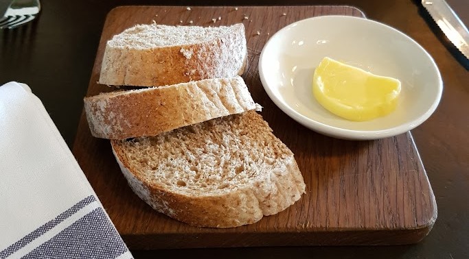 
Three slices of rustic, wholegrain bread served on a wooden board, accompanied by a small white dish containing a pat of creamy butter. A neatly folded white and blue striped napkin is placed alongside, adding a touch of simple elegance to this traditional presentation of bread and butter, ready to be enjoyed as part of a meal.