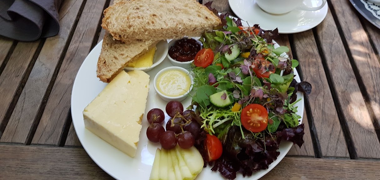 A classic Ploughman's lunch served on a white plate featuring thick slices of rustic brown bread and butter, a large wedge of cheddar cheese, fresh salad with mixed greens, cherry tomatoes, cucumbers, and colorful peppers. Accompanying the meal are a small bunch of grapes, slices of apple, and a pot of chutney. A cup of tea sits beside the plate, completing the traditional English dining experience. The meal is laid out on a wooden table in a charming outdoor setting.
