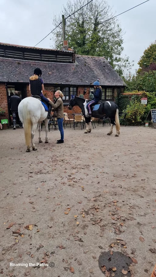 
This image captures a charming outdoor scene in front of a rustic brick building, likely part of a countryside café or farm shop. Two riders on horseback are stopped outside, with one woman interacting with the riders, possibly adjusting tack or giving instructions.

The horses, one white and the other black and white, stand calmly as they wait, and both riders are wearing helmets and casual riding attire. The ground is scattered with dry leaves, adding an autumnal touch. The building in the background has a tiled roof, ivy-covered walls, and an inviting setup with tables and chairs, suggesting it’s a popular stop for both locals and passersby, including those on horseback. This peaceful scene reflects the leisurely pace and close-to-nature lifestyle typical of rural settings.