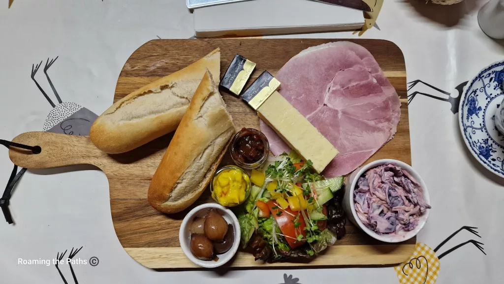 
This image shows a beautifully arranged Ploughman’s Lunch at The Old Forge, displayed on a wooden serving board. The classic British meal includes:

Two baguette halves – Fresh and crusty, perfect for pairing with the cheese and ham.
Thick slice of cheddar cheese – A creamy, yellow wedge of cheddar, a traditional element of the Ploughman’s.
Sliced ham – Generous, thin slices of pink ham, offering a mild, savory flavor.
Condiments and accompaniments:
A small jar of chutney, likely caramelized onion or fruit-based, for a sweet and tangy kick.
A small jar of piccalilli – a bright yellow relish made from pickled vegetables in a mustard sauce, adding a sharp, tangy note.
A small bowl of pickled onions, adding a sour punch.
Mixed side salad – Fresh lettuce, tomatoes, cucumber, and yellow bell pepper with microgreens, bringing vibrant color and crunch.
Creamy coleslaw – A bowl of pinkish coleslaw with red cabbage, adding a creamy and crunchy texture.
The board rests on a farm-themed tablecloth featuring playful chicken illustrations, enhancing the rustic and homely presentation of this classic Ploughman’s Lunch.