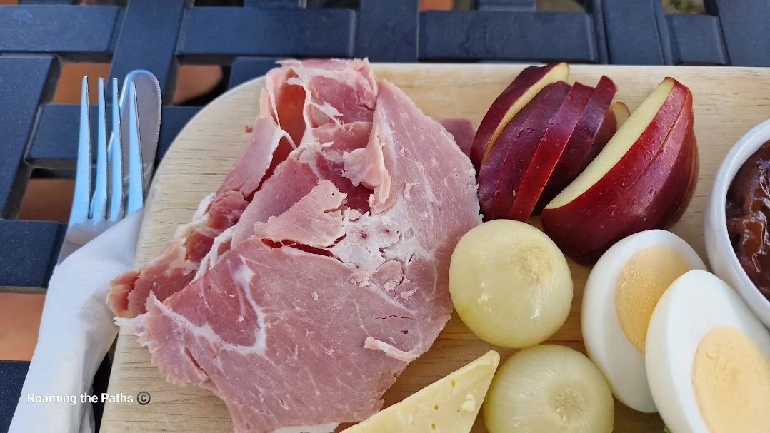 Close-up of a wooden platter featuring thick slices of rustic ham, sliced red apple, pickled onions, and halved boiled eggs. A wedge of cheese and a glimpse of chutney in a ramekin are visible on the side. A napkin-wrapped knife and fork rest on the edge of the board, all set atop a black metal outdoor table.