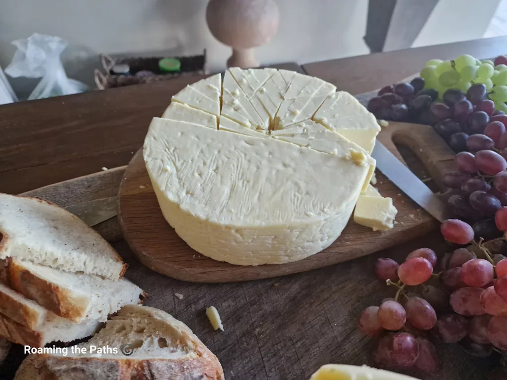 A rustic wooden board set with a round wheel of pale yellow cheese, neatly cut into wedges, surrounded by slices of crusty bread and bunches of fresh red and green grapes. The setting evokes a homely, countryside feel, perfect for a traditional Ploughman’s-style spread.