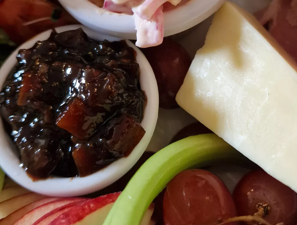 Close-up of a small white ramekin filled with chunky dark brown pickle beside a wedge of cheese, red grapes, celery, and apple slices on a Ploughman’s lunch plate.