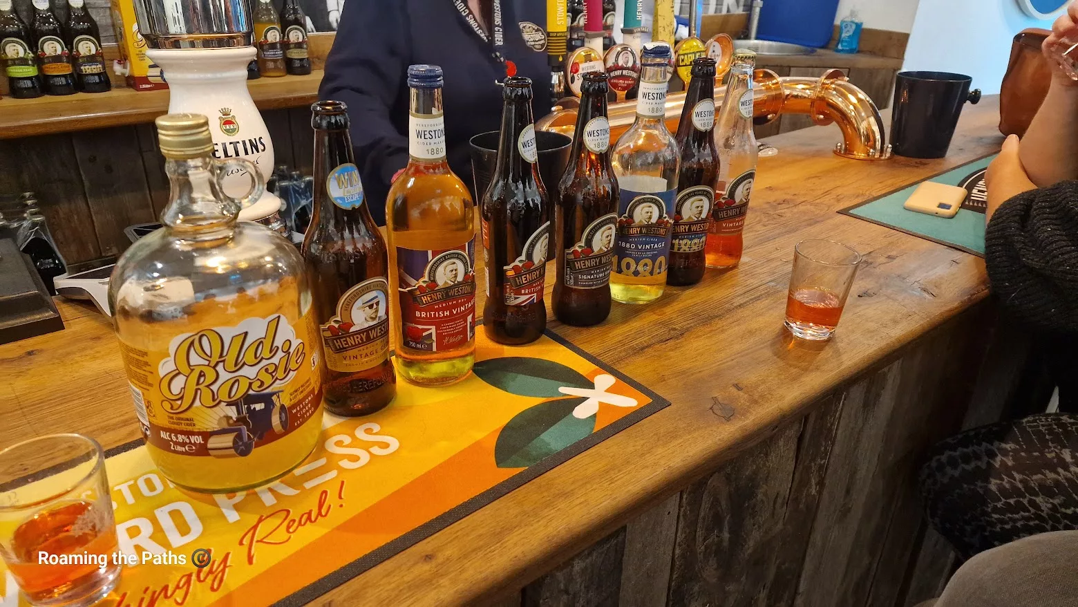 A bar counter displaying a selection of Westons ciders, including Old Rosie and several Henry Westons varieties, arranged in a line for tasting. Two small glasses with cider samples sit on the wooden bar beside the bottles, with people chatting nearby.