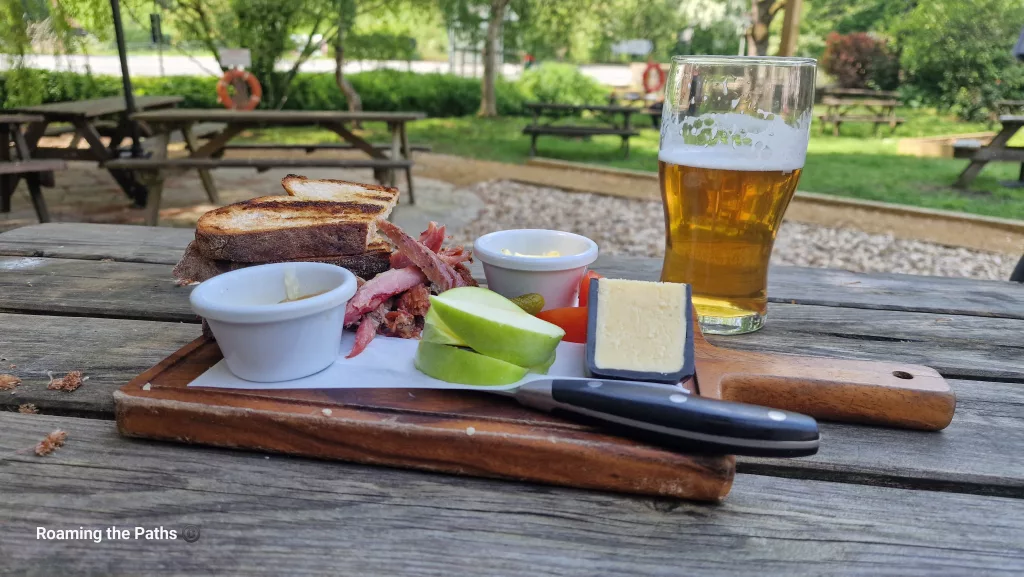 Traditional Ploughman’s lunch with cheese, ham, toasted bread, apple slices, pickles, and a pint of beer served on a wooden board in a sunny pub beer garden.
