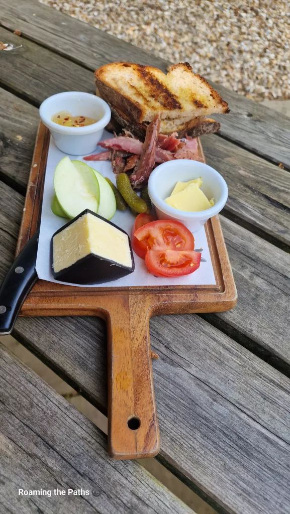 Traditional Ploughman’s lunch served on a wooden paddle board with toasted bread, carved ham, mature cheese, apple slices, pickles, tomato, butter, and chutney on a rustic picnic table.