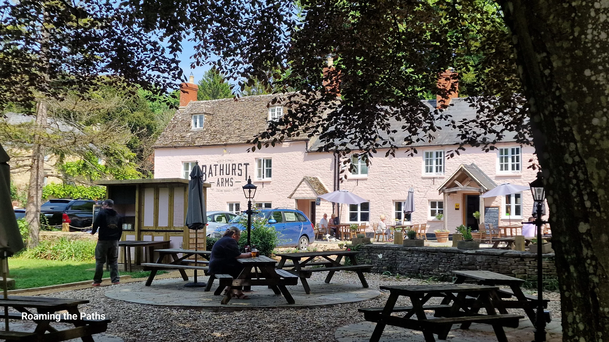 The Bathurst Arms pub at North Cerney viewed from a shaded beer garden, with picnic benches, mature trees, and visitors enjoying a sunny afternoon outside the traditional Cotswold inn.