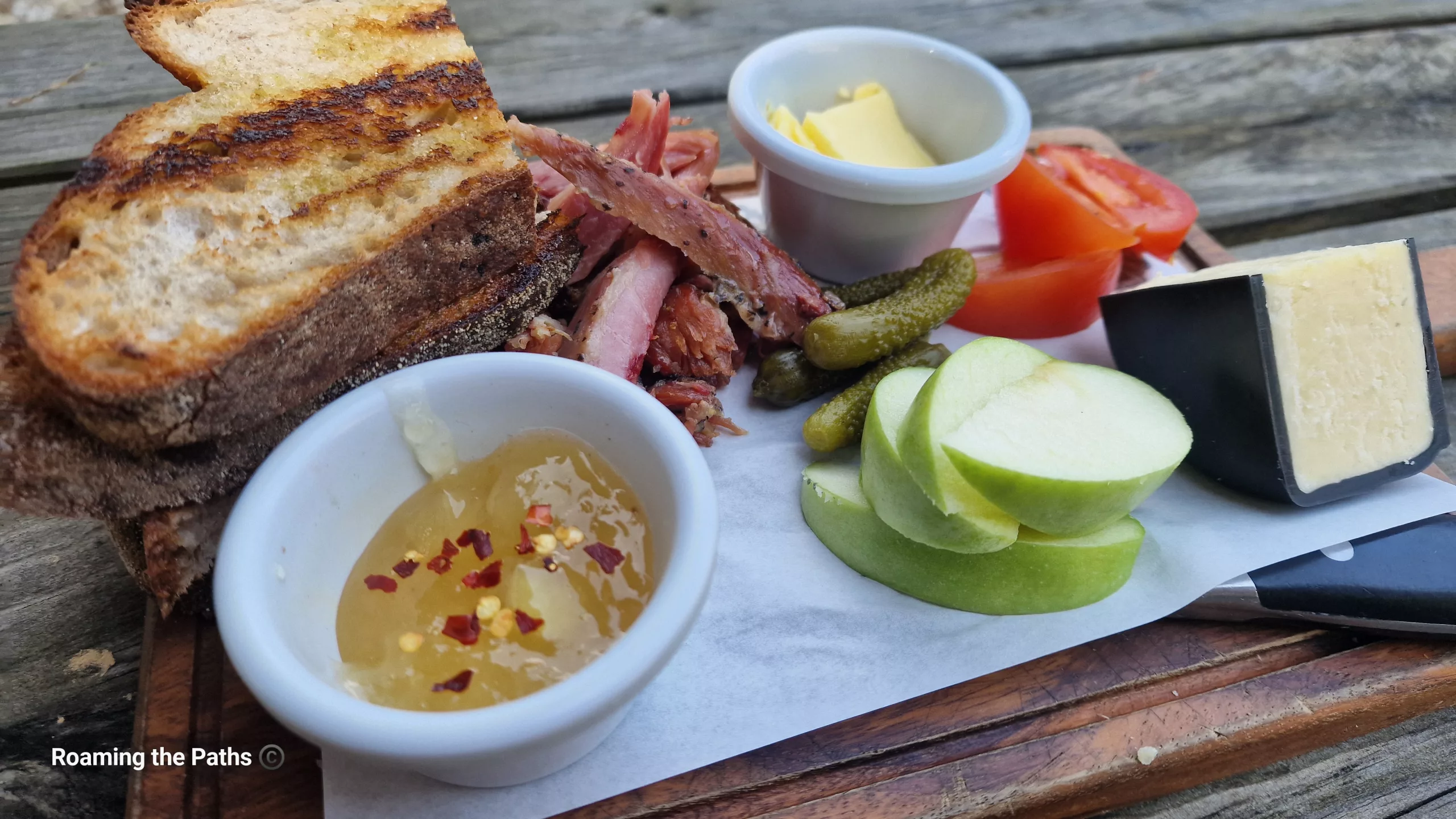 Close-up of a rustic Ploughman’s lunch on a wooden serving board, featuring toasted farmhouse bread, carved ham, mature cheese, sliced green apple, pickles, tomato wedges, butter, and a small pot of chutney, served outdoors on a picnic table.
