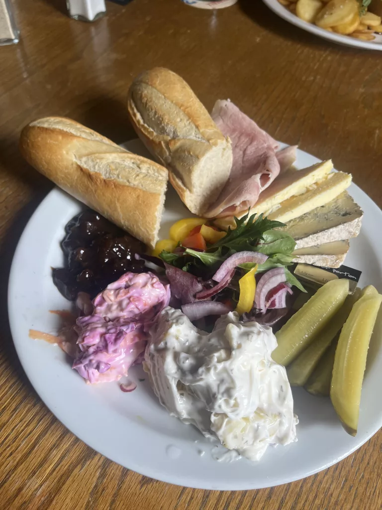 A pub-style Ploughman’s lunch served on a white plate on a wooden table. The meal includes crusty baguette, slices of ham, several cheeses including blue cheese, pickled gherkins, mixed salad leaves with red onion and peppers, creamy potato salad, pink coleslaw, and Branston Pickle.
