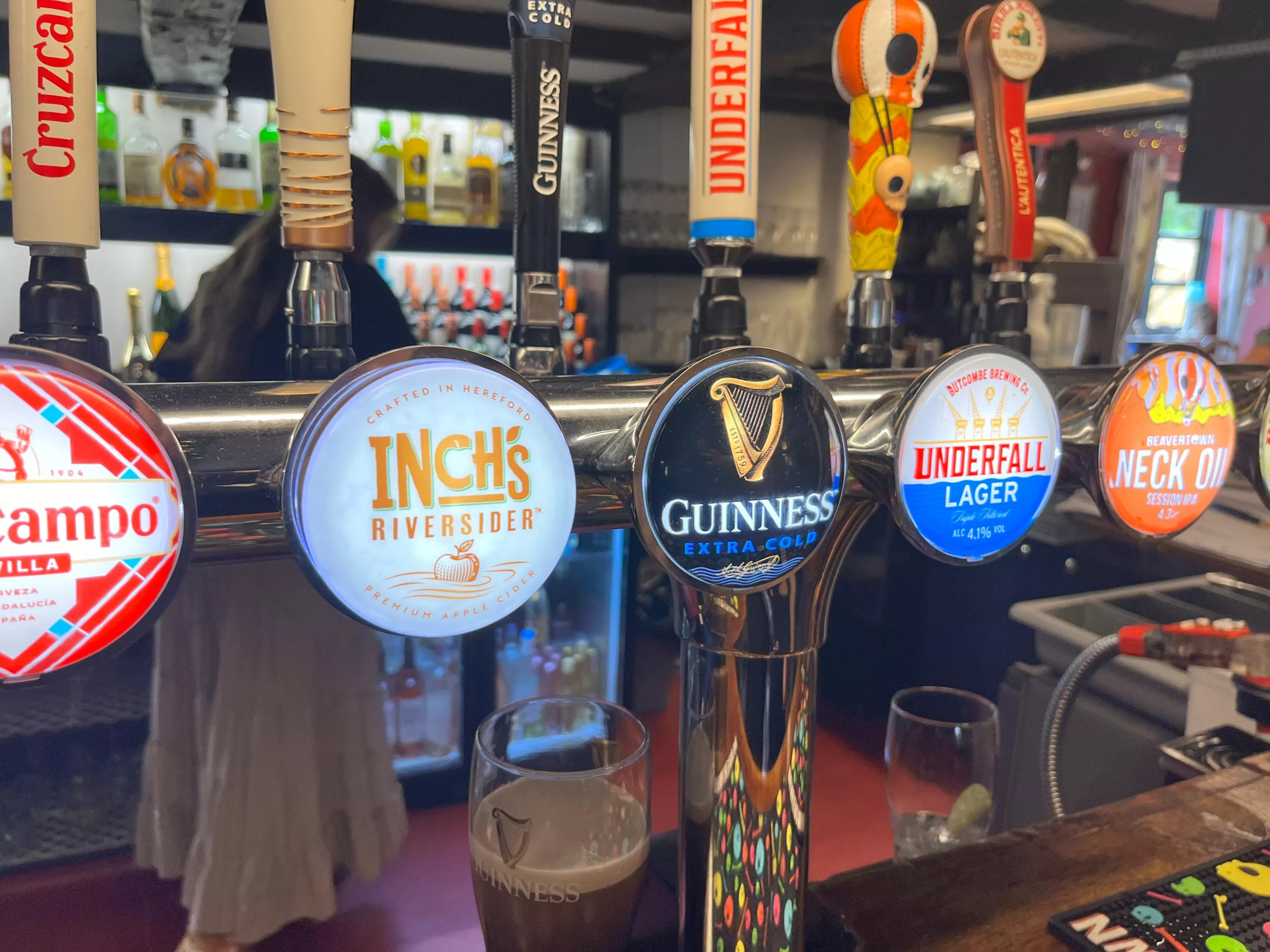 A row of beer taps behind a pub bar, featuring Cruzcampo, Inch’s Cider, Guinness Extra Cold, Underfall Lager, and Beavertown Neck Oil. Bottles and glasses are visible in the softly lit background, with a partly poured pint of Guinness in the foreground.