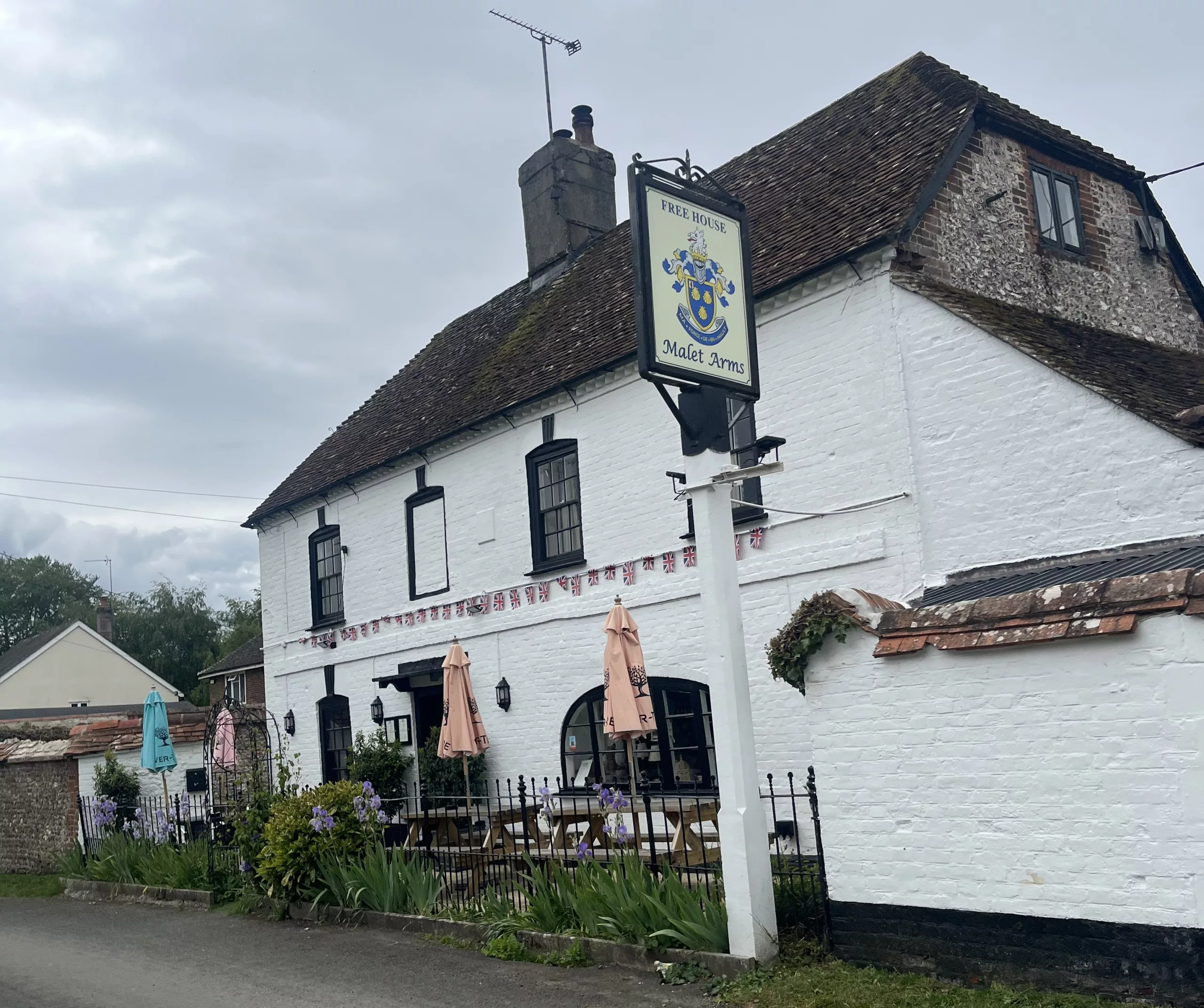 The exterior of the Malet Arms, a traditional white-painted country pub with black trim and a steep tiled roof. A hanging pub sign stands beside the building, with outdoor seating, umbrellas, flowers, and bunting decorating the frontage beneath a cloudy sky.