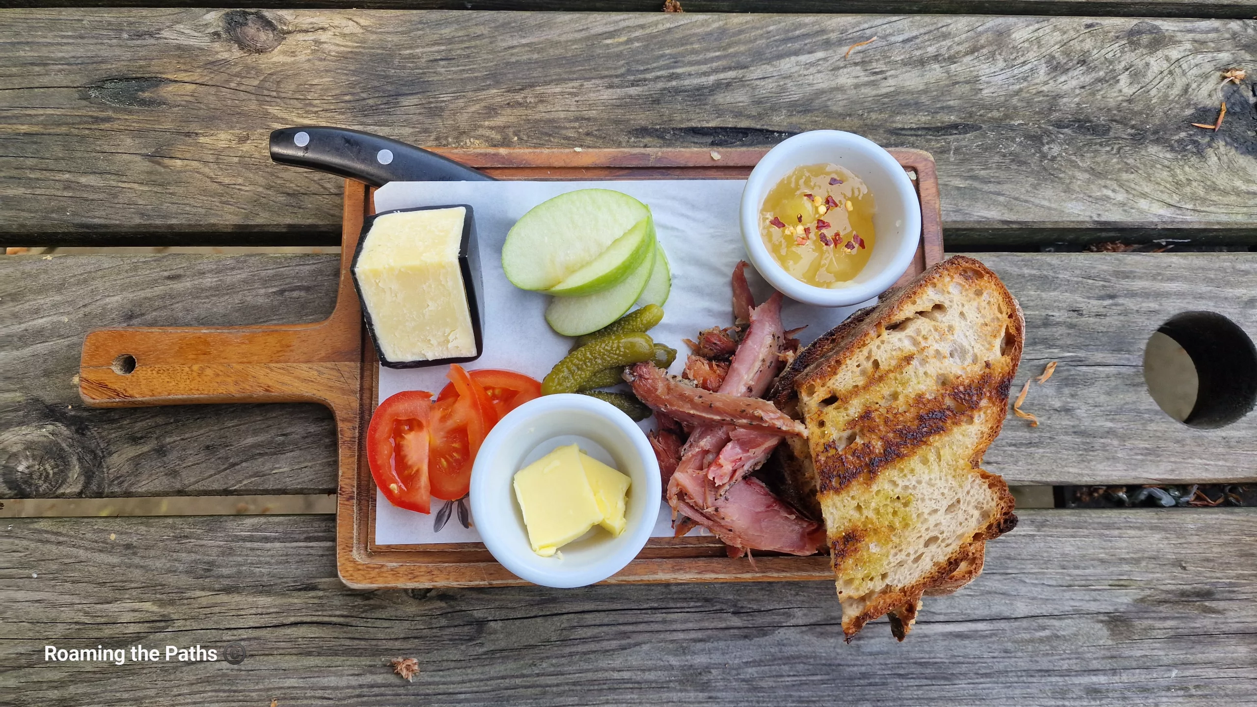 Overhead view of a rustic Ploughman’s lunch on a wooden serving board with toasted bread, ham, mature cheese, apple slices, pickles, tomato, butter, and chutney on a weathered picnic table.