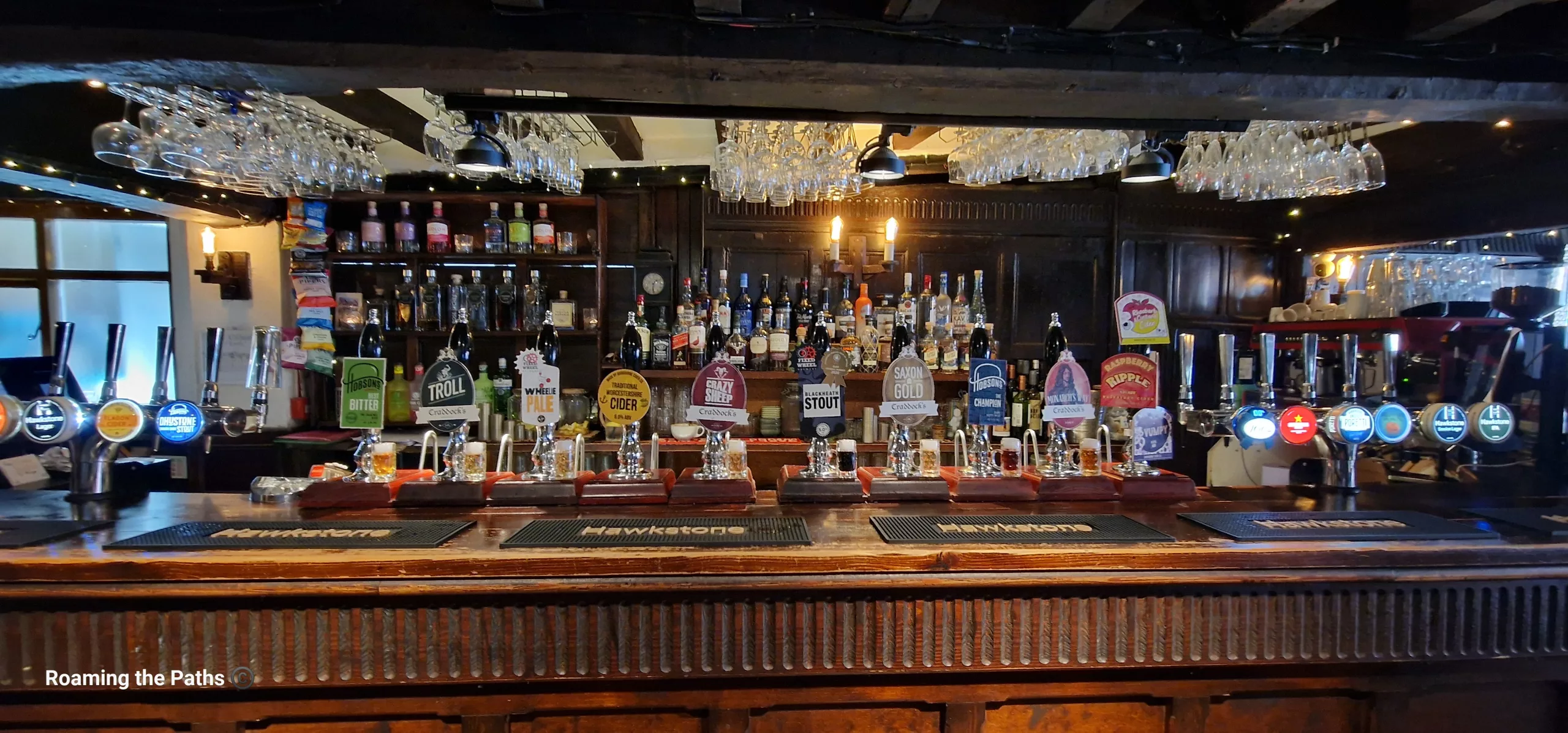 The traditional bar of an English pub, featuring a long row of hand-pulled ales, ciders, and craft beers. Hanging glassware, dark wood panelling, and shelves stocked with spirits create a warm and inviting atmosphere behind the polished wooden counter.