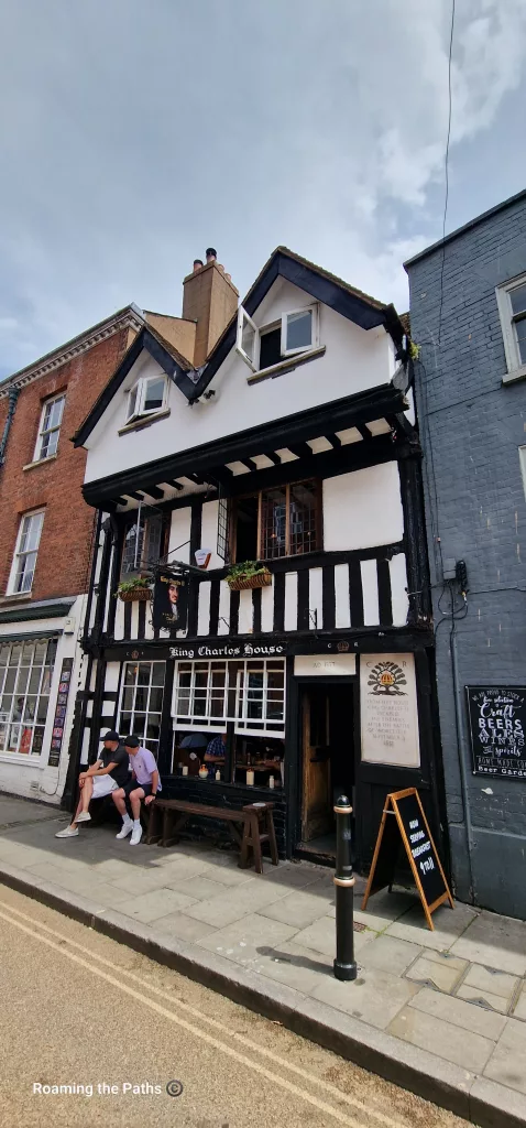 The historic King Charles House pub in Worcester, a striking black-and-white timber-framed building dating from the sixteenth century. Hanging baskets decorate the upper windows, while visitors sit outside on a bench beneath the traditional pub sign.
