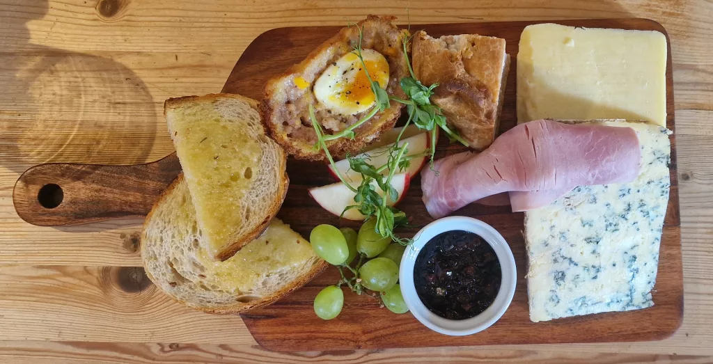 Overhead view of a traditional Ploughman’s lunch served on a wooden paddle board. The meal includes two cheeses – a large wedge of blue cheese and a mature hard cheese – alongside slices of ham, a Scotch egg, a pork pie, crusty buttered bread, grapes, apple slices, pea shoots, and a pot of dark chutney. The board is presented on a rustic wooden table.
