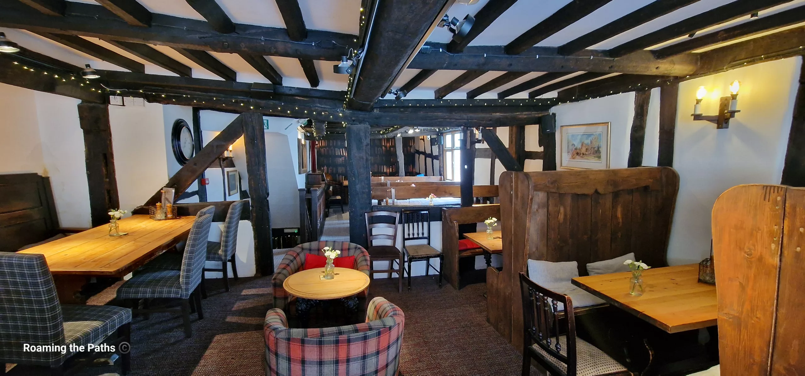 A cosy dining area inside a historic timber-framed pub. Exposed black oak beams, wooden booths, tartan armchairs, and polished wooden tables create an inviting atmosphere, with small flower vases and soft lighting adding to the traditional character.