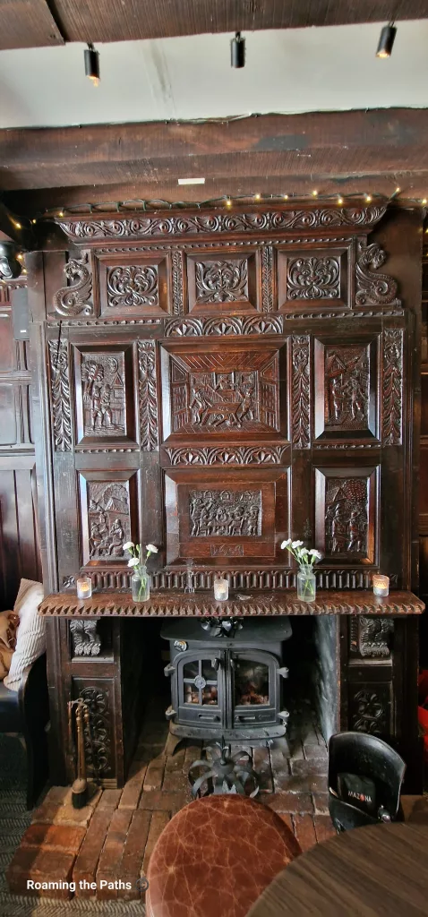 An ornate carved oak fireplace inside a historic pub. Intricate wood panelling decorated with figures, scrollwork, and decorative motifs surrounds a cast-iron wood-burning stove. Small vases of flowers and candles sit on the mantel shelf, adding warmth to the traditional interior.
