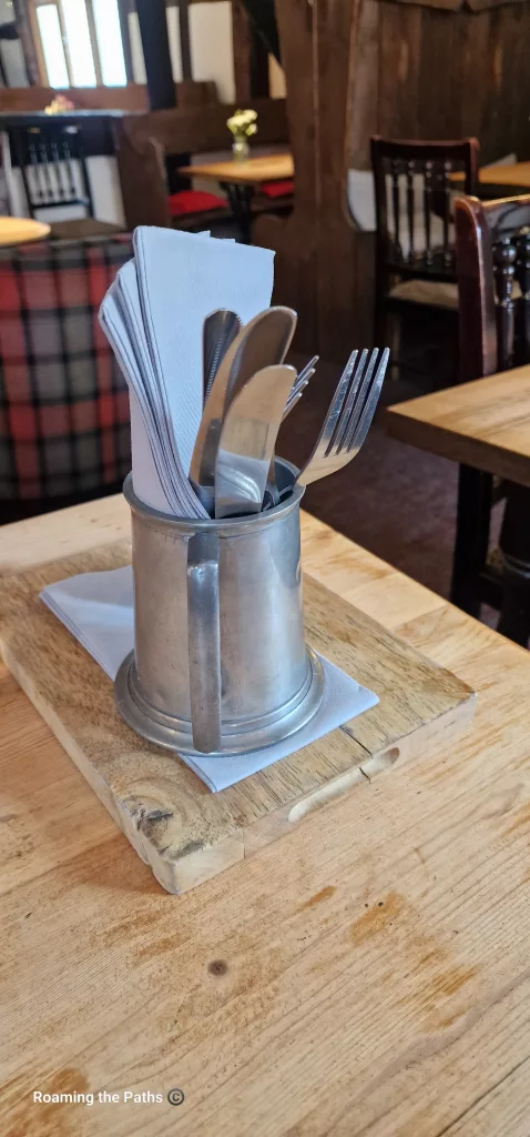 A pewter-style tankard holding cutlery and folded napkins sits on a wooden serving board atop a rustic timber table. In the background, wooden booths, tartan seating, and exposed beams create the atmosphere of a traditional English pub.